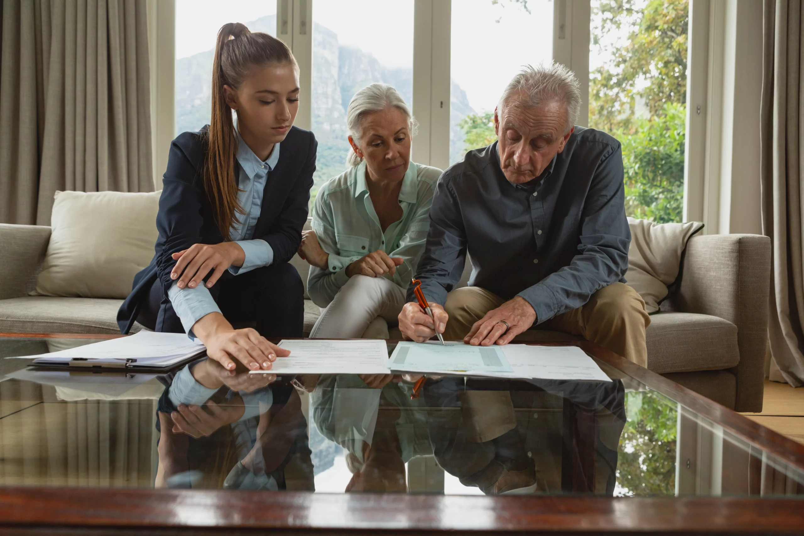 Elderly couple reviews documents with a young businesswoman on a sofa. The man signs papers, conveying a serious, focused atmosphere.