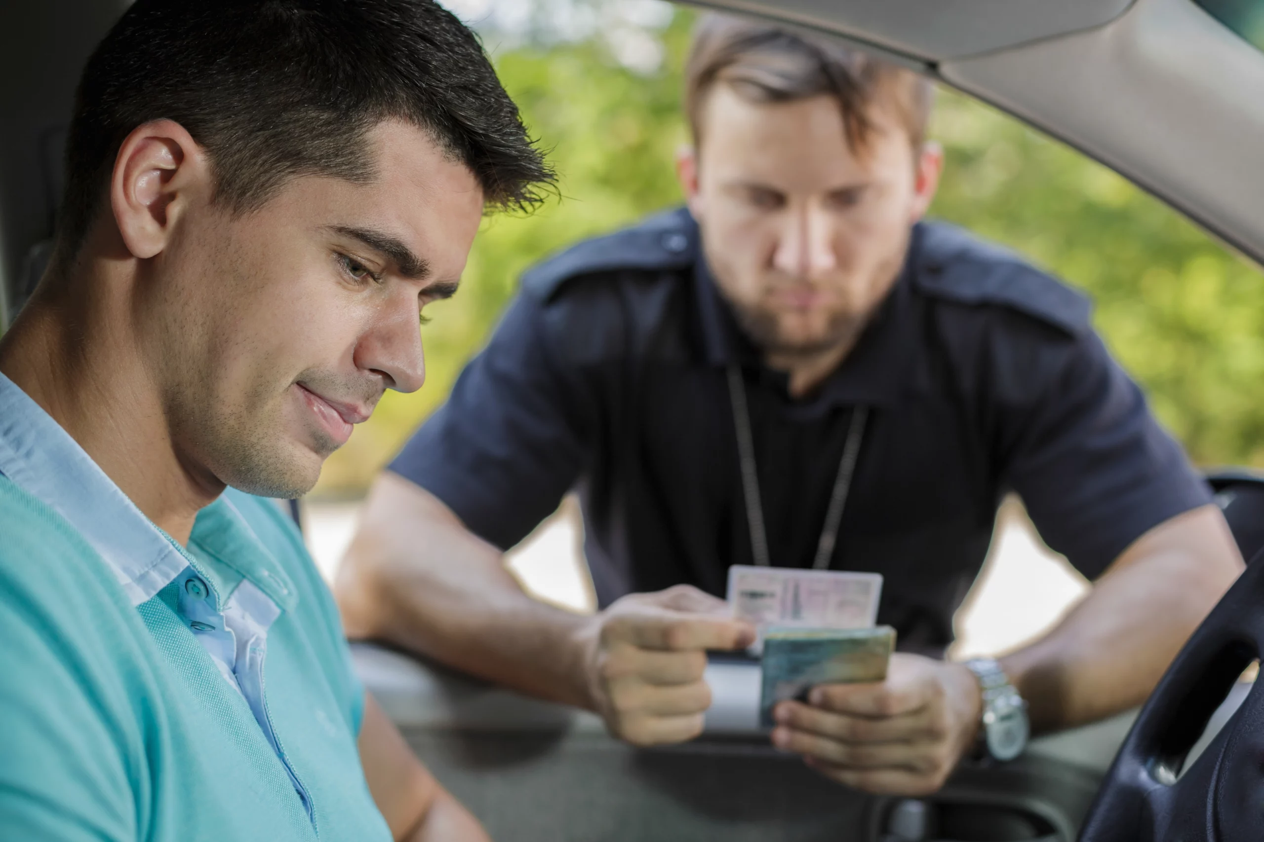 A driver in a car hands his license to a police officer standing outside. The officer examines the document, conveying a serious yet routine mood.