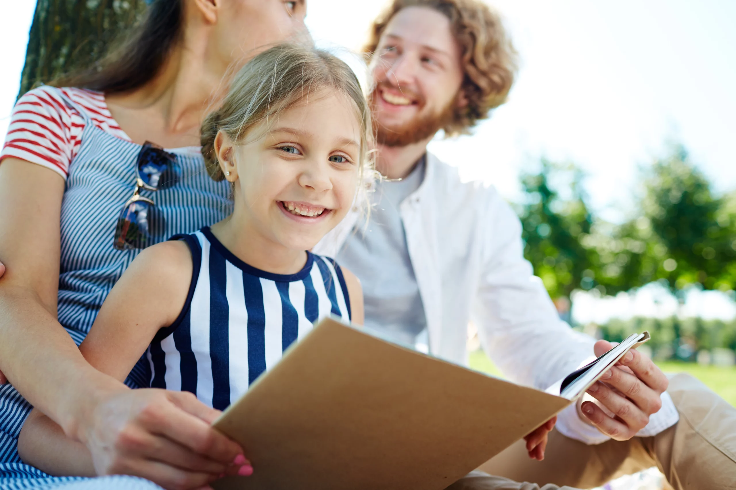 Smiling young girl in a striped dress holds a book while sitting with two adults under a tree in a bright green park — symbolizing family, love and planning for the future.