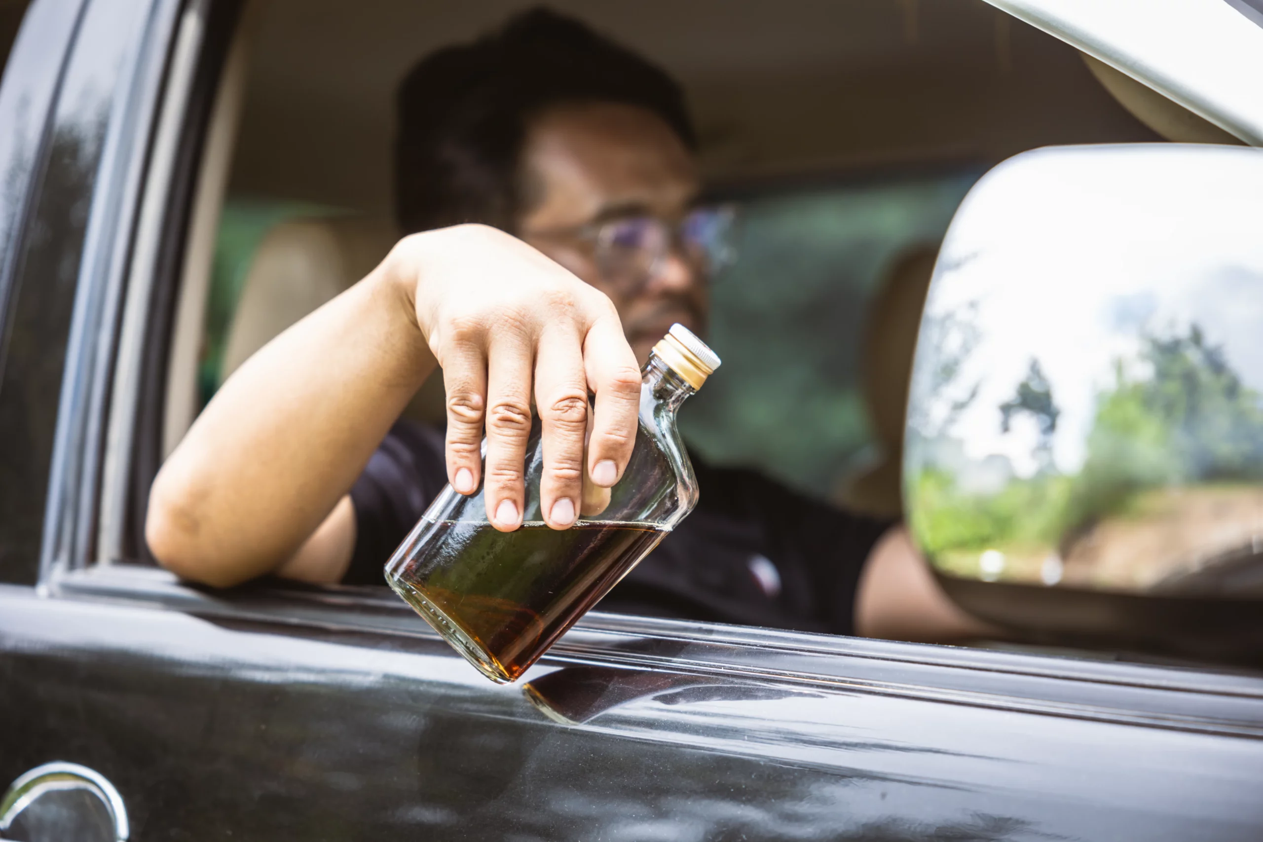 Driver holding a liquor bottle out of a car window, illustrating risky DUI behavior and the serious legal consequences that can stay on a California driving record for up to 10 years.