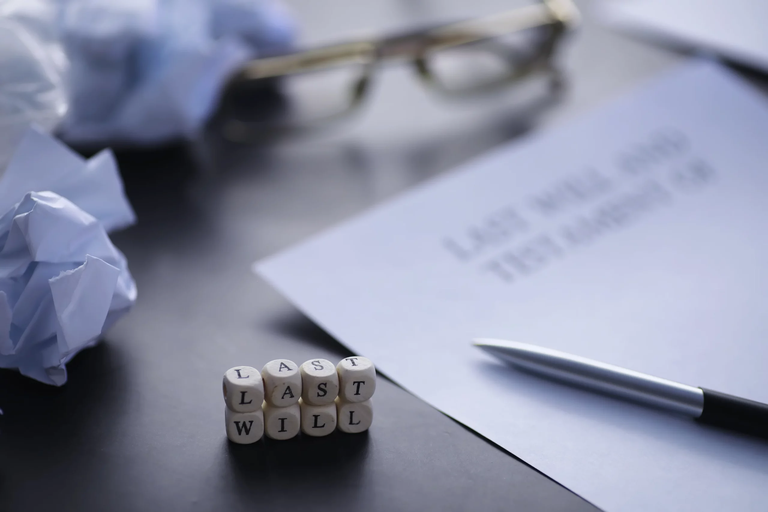 Wooden blocks spelling ‘Last Will’ beside a signed will document, pen, and glasses, representing estate planning in California and the need for both a will and a living trust to properly manage and transfer assets.