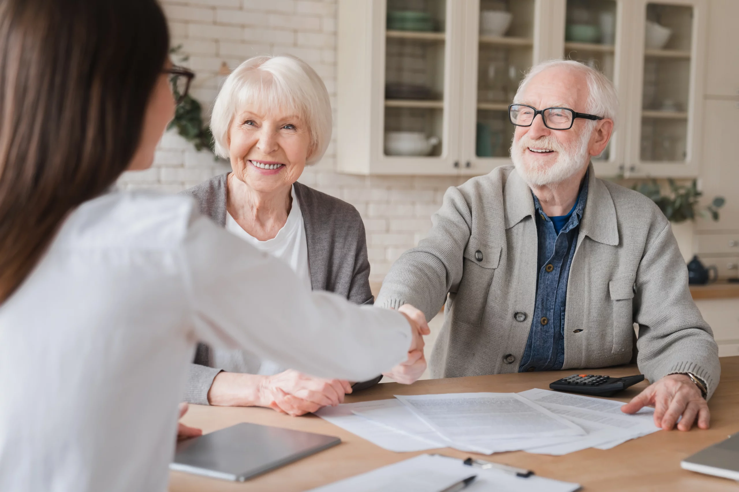 Elderly couple smiling and shaking hands with an advisor over estate planning documents, representing California guidance on using both a living trust and a will to properly manage and transfer assets.