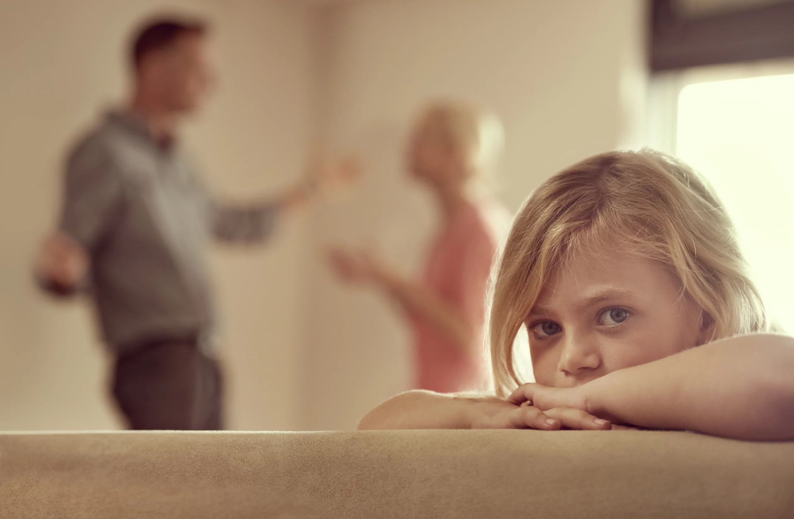Sad child sitting alone while parents argue in the background, representing the emotional impact when a parent refuses to follow a court-ordered parenting plan in California custody cases.