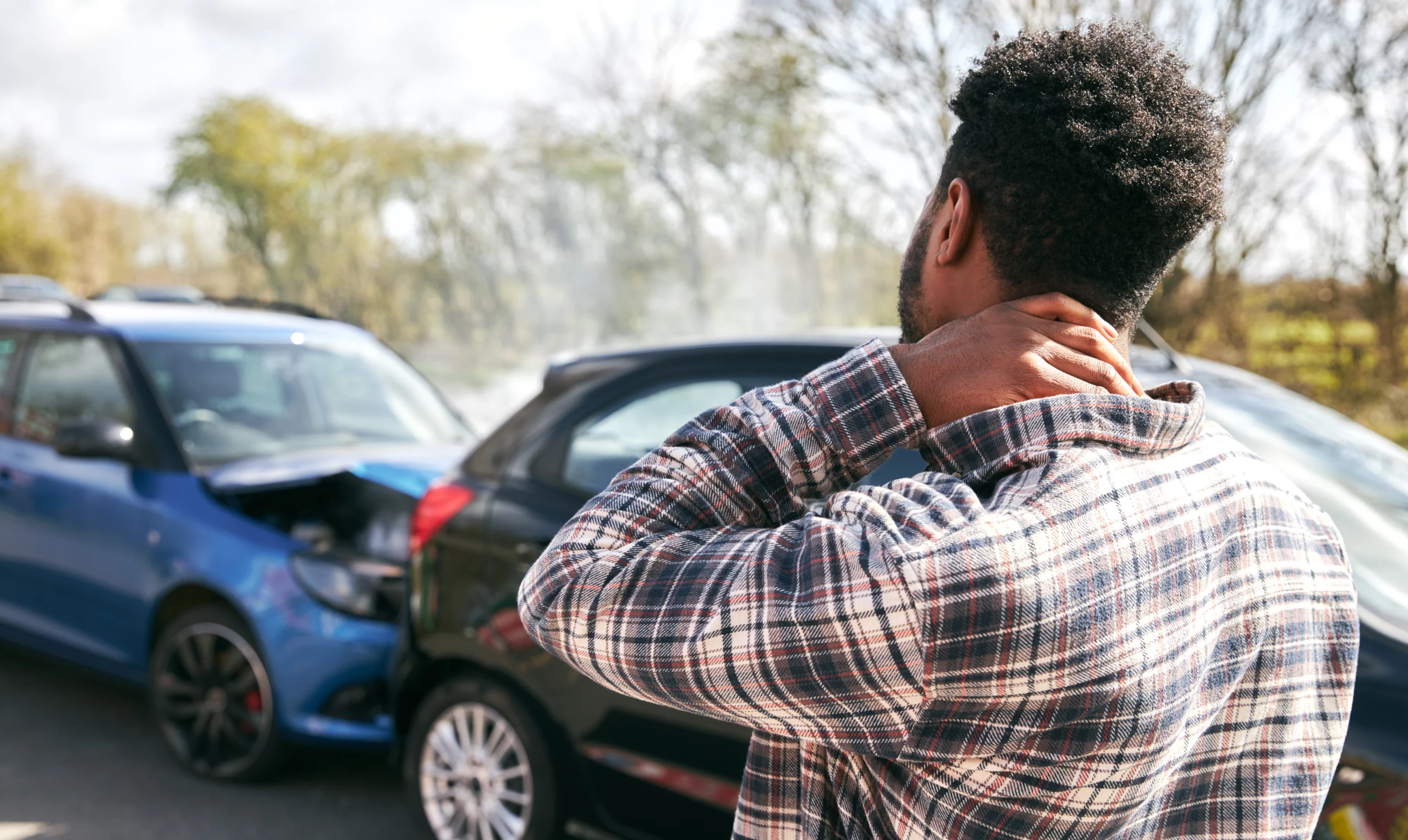 Man in a plaid shirt rubbing his neck after a car accident between a blue and black vehicle, illustrating what to do if the other driver has no insurance in California and steps to protect your personal injury claim.