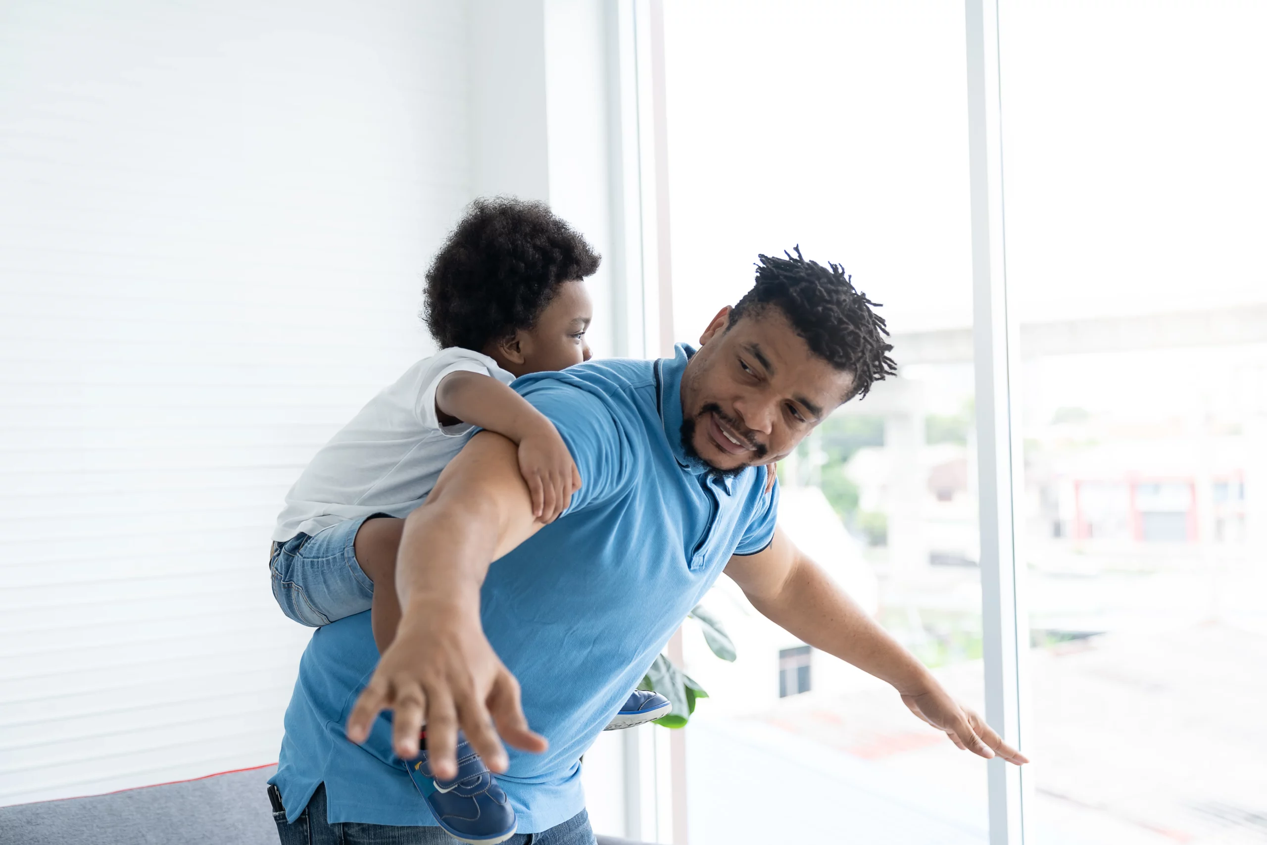 Father in blue polo shirt playfully carrying smiling child on his back in sunlit room, representing fathers seeking full custody and strong parent‑child bond under California family law