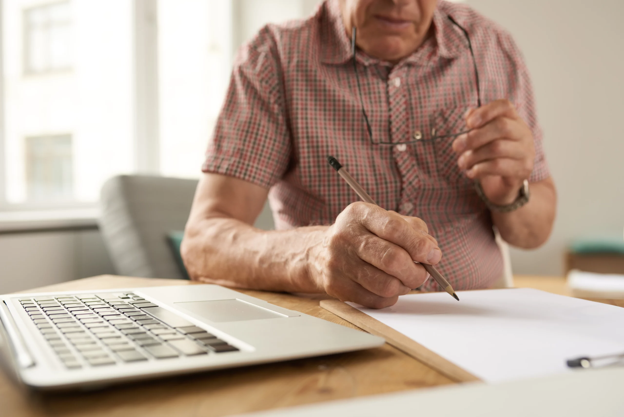 Elderly man in plaid shirt holding glasses and writing on paper next to a laptop in sunlit room, representing a senior preparing a simple will and considering legal help in California estate planning